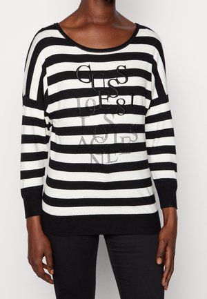 Sort-hvid, vandret stribet sweater med teksten "GUESS 1981" på fronten, båret med sorte bukser.