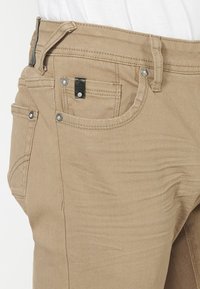 Pantalones de algodón beige con corte estándar, cinco bolsillos, costuras reforzadas y detalles en metal.