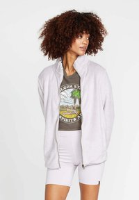 Giacca in pile grigia chiara con zip frontale, collo alto e texture morbida. Indossata sopra una t-shirt con grafica e abbinata a pantaloni corti leggeri.