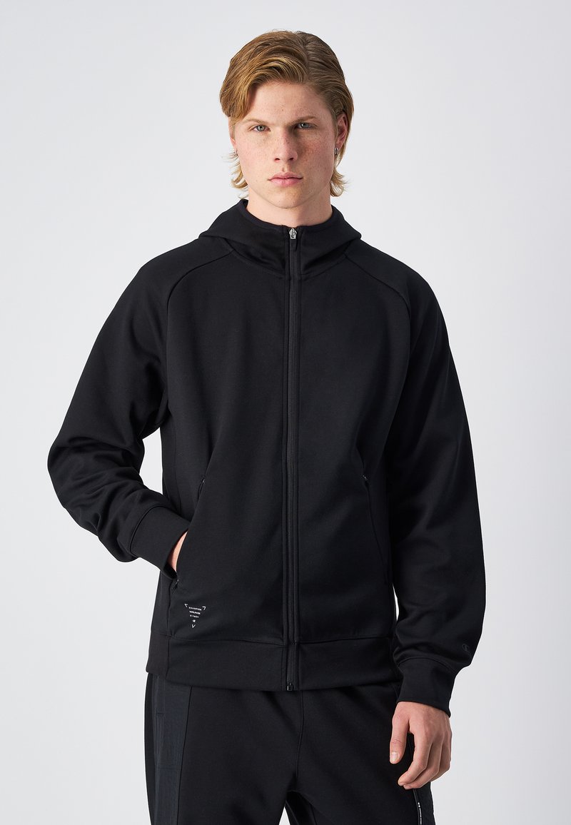 Sudadera negra con cremallera, fabricada en tejido suave. Presenta un cuello alto, dos bolsillos laterales y un pequeño logotipo cerca del dobladillo. Diseño simple y atlético.