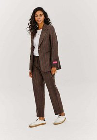 Hnědý pruhovaný oblek z texturovaného materiálu, který zahrnuje přizpůsobenou blazer s knoflíkem, sladěné kalhoty a bílé tenisky.