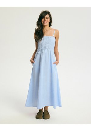 Kobieta uśmiechająca się, ubrana w jasnoniebieską, bezrękawną sukienkę maxi w kratę z kieszeniami i brązowe buty wsuwane, stojąca na prostym jasnym tle.