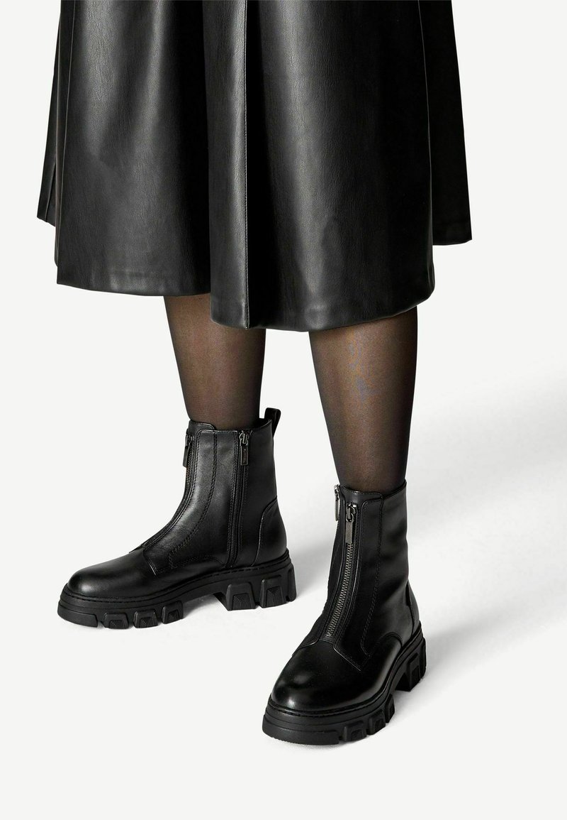 Bottes en cuir noires mi-mollet avec semelles épaisses et fermetures éclair frontales, portées avec des collants noirs transparents et une jupe en cuir noire.