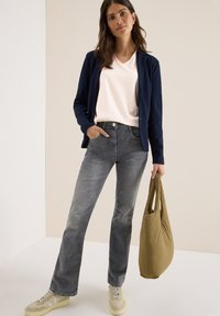Marineblazer over een lichtroze V-hals shirt, gecombineerd met grijze flared jeans en beige sneakers, en een bruine tote bag vast.