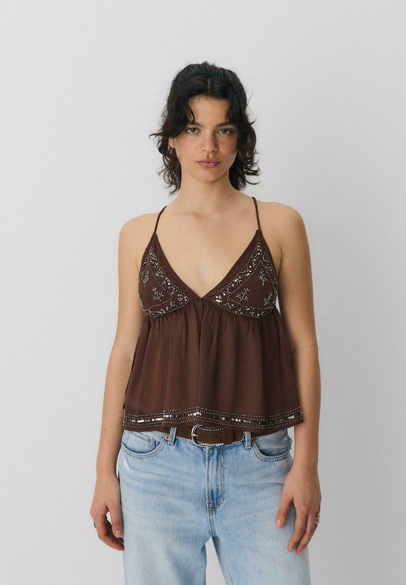 Femme portant un haut marron brodé sans manches avec de fines bretelles et un jean bleu clair sur un fond gris clair uni.