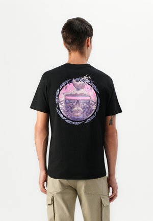 Man in een zwart T-shirt met een paarse schedel, vlinder en een cirkelvormig tekstontwerp op de achterkant, gecombineerd met beige cargobroeken.