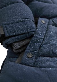 Dunkelblauer Steppjacken-Detail mit Wassertropfen, ausgestattet mit strukturiertem Stoff, schwarzen Knopfakzenten und einem hervorgehenden Strickbündchen.