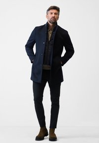 Marinblå rock med hög krage och knappstängning, lager på lager över en svart väst med dragkedja. Slimmade svarta byxor och bruna mockastövlar till ankeln.