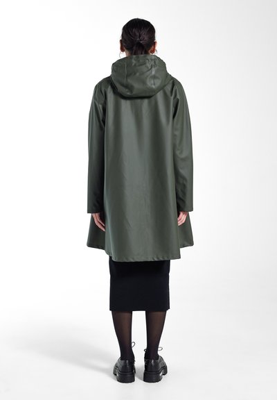 Manteau imperméable vert avec capuche, présentant une coupe ample, des manches longues et une texture lisse, associé à une jupe noire et des chaussures chunky.