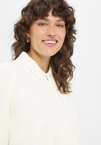 Licht crèmekleurig topje met een kraag, een klein zilveren accent en een gestructureerde stof, met een open zak aan de zijkant.