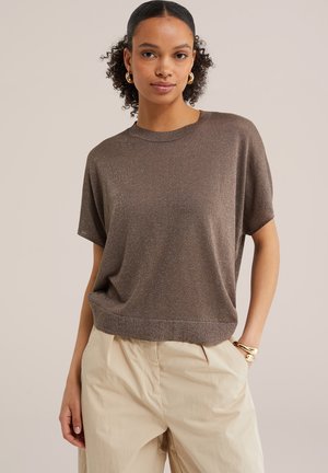 Nő rövid ujjú barna kötött felsőt és bézs nadrágot visel, arany karika fülbevalóval és arany karkötővel kiegészítve.