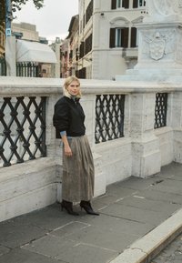 Mladá žena s blond vlasmi, oblečená v čiernej bunde, dlhej hnedej sukni a čiernych topánkach, opierajúca sa o kamenné zábradlie v európskom meste.