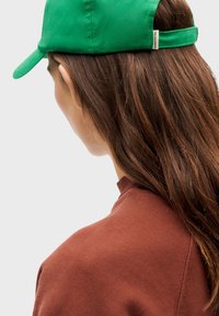 Groene pet van gladde stof met een gebogen rand; gedragen met een bruine, gestructureerde trui. Haar valt over de schouder en toont een kleurcontrast.