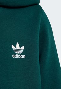 Sweat à capuche vert en tissu doux, avec un logo Adidas brodé blanc comportant trois bandes et un motif de feuille sur la partie gauche de la poitrine.