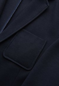 Gros plan sur la poche poitrine et le revers d'un blazer bleu marine, montrant un tissu texturé et des coutures soignées.