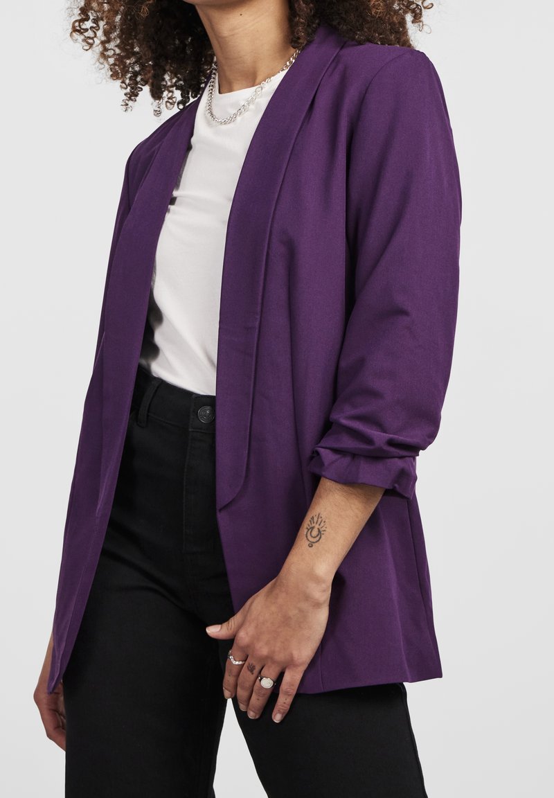 Femme portant un blazer violet sur une chemise blanche et un pantalon noir, montrant un collier argenté, des bagues et un tatouage au poignet représentant un croissant de lune et des rayons de soleil.