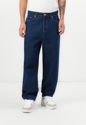 Jean en denim bleu foncé avec une coupe droite, doté d'une fermeture à bouton, de passants de ceinture et d'un design classique à cinq poches. Porté avec des baskets blanches.