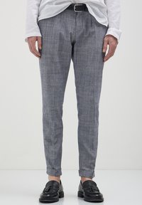 Uomo che indossa pantaloni grigi skinny, cintura di pelle nera, mocassini neri con nappine e una camicia bianca a maniche lunghe infilata su uno sfondo semplice.