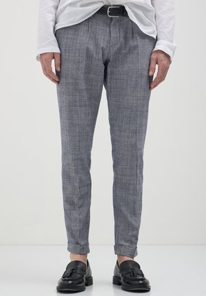 Homme portant un pantalon gris ajusté, une ceinture en cuir noire, des mocassins à glands noirs et une chemise blanche à manches longues rentrée sur un fond uni.