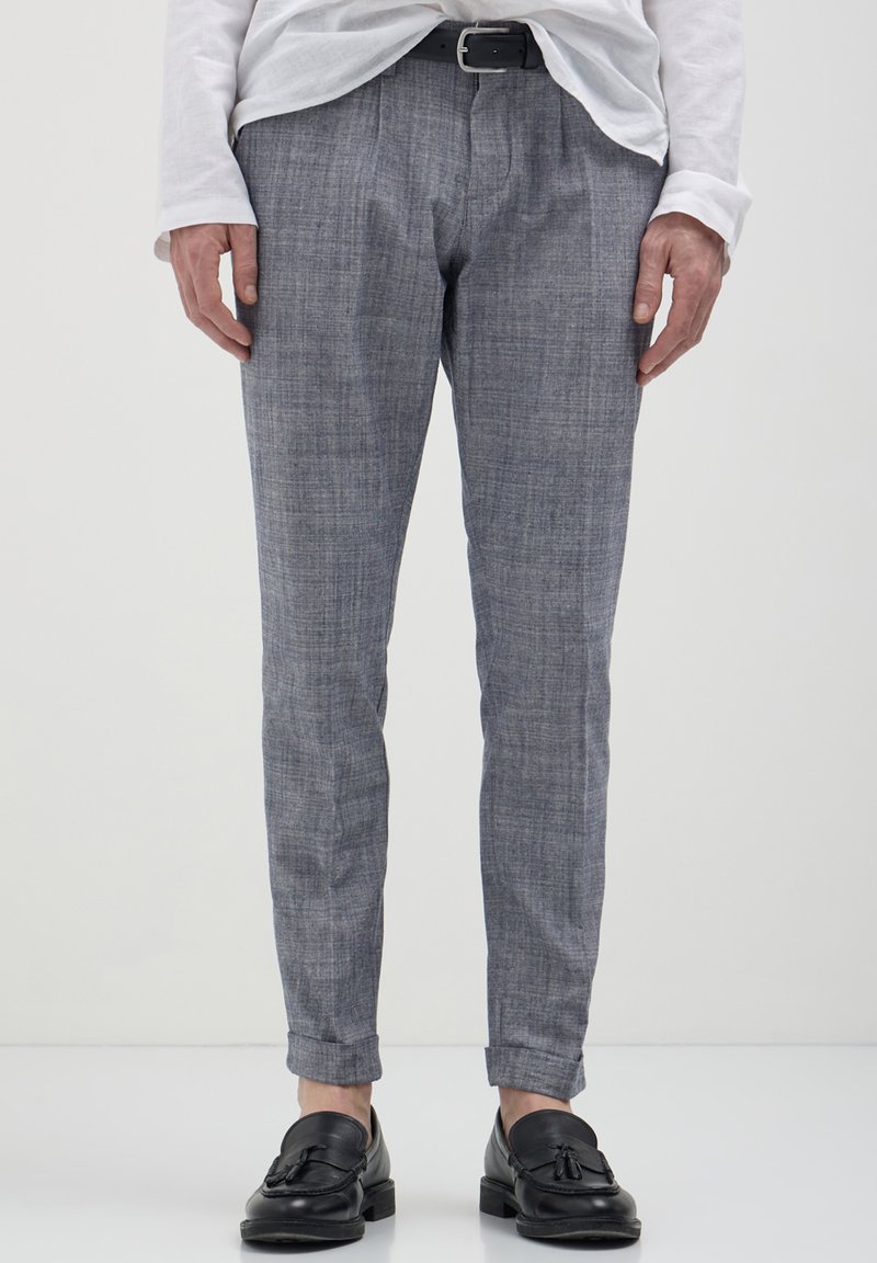 Uomo che indossa pantaloni grigi skinny, cintura di pelle nera, mocassini neri con nappine e una camicia bianca a maniche lunghe infilata su uno sfondo semplice.