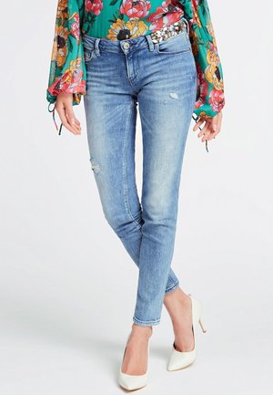 Frau trägt hellblaue, zerrissene Skinny-Jeans kombiniert mit weißen High Heels und einer grünen, geblümten Langarmbluse mit Schmuckdetail an der Taille.
