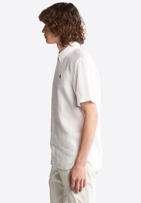Camisa de lino blanca de manga corta con cuello abotonado, que presenta un sutil logo bordado en el pecho y un corte relajado.