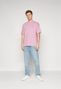 Lyserød bomulds t-shirt med korte ærmer og en afslappet pasform, med små sorte bogstaver på brystet, parret med lyseblå jeans og hvide sko.