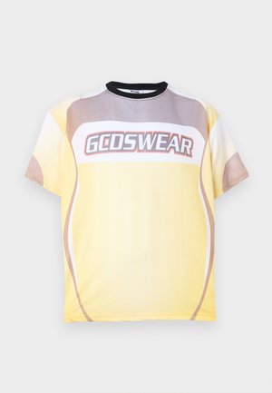 Homme debout, une main dans la poche, portant un t-shirt GODSWEAR jaune, beige et blanc, pantalon noir et chaussures noires sur un fond gris clair.