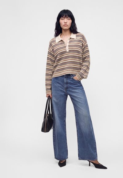 Femme aux cheveux noirs portant un polo à manches longues rayé, un jean bleu à jambes larges, des talons noirs et tenant un sac à main noir.