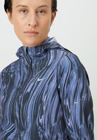 Zbliżenie osoby w niebieskiej i czarnej kurtce Nike z zamkiem błyskawicznym i kapturem na tle białej ściany.