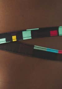 Una fascia di tessuto multicolore presenta un motivo geometrico con elementi turchesi, gialli, rossi e neri, contrastati su uno sfondo marrone.