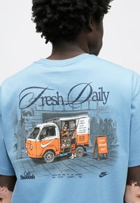 Gaiši zils kokvilnas T-krekls ar grafisku attēlu — kafijas ratiņš ar oranžiem akcentiem, cilvēki rindā un teksts, kas veicina "Svaigs katru dienu" un "Cafe Swoosh".