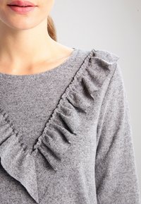 Femme portant un pull gris avec des détails en tissu volanté le long de l'encolure en V, montrée des épaules aux lèvres.