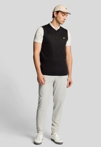 Maglione nero a maglia senza maniche sopra una camicia bianca con colletto, abbinato a pantaloni grigio chiaro e sneakers bianche. Cappello beige con accentuazione del logo.