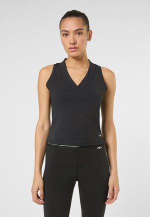 Frau trägt ärmelloses schwarzes V-Ausschnitt-Sporttop und schwarze Leggings mit FILA-Logo auf beiden, steht vor einem einfarbigen Hintergrund.