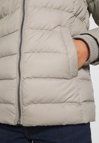 Personne portant une veste matelassée beige clair avec fermeture éclair devant, main partiellement dans la poche latérale, bord de pull gris visible et jean bleu foncé.
