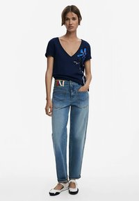 Marineblå v-hals t-shirt med blå blomstret broderi, kombineret med lyseblå løstsiddende jeans og hvide og sorte slip-on sko.