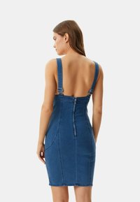 Denim dress with wide shoulder straps, back zipper, fitted design, and backless style. Dark blue color, smooth texture, and angled stitching details.