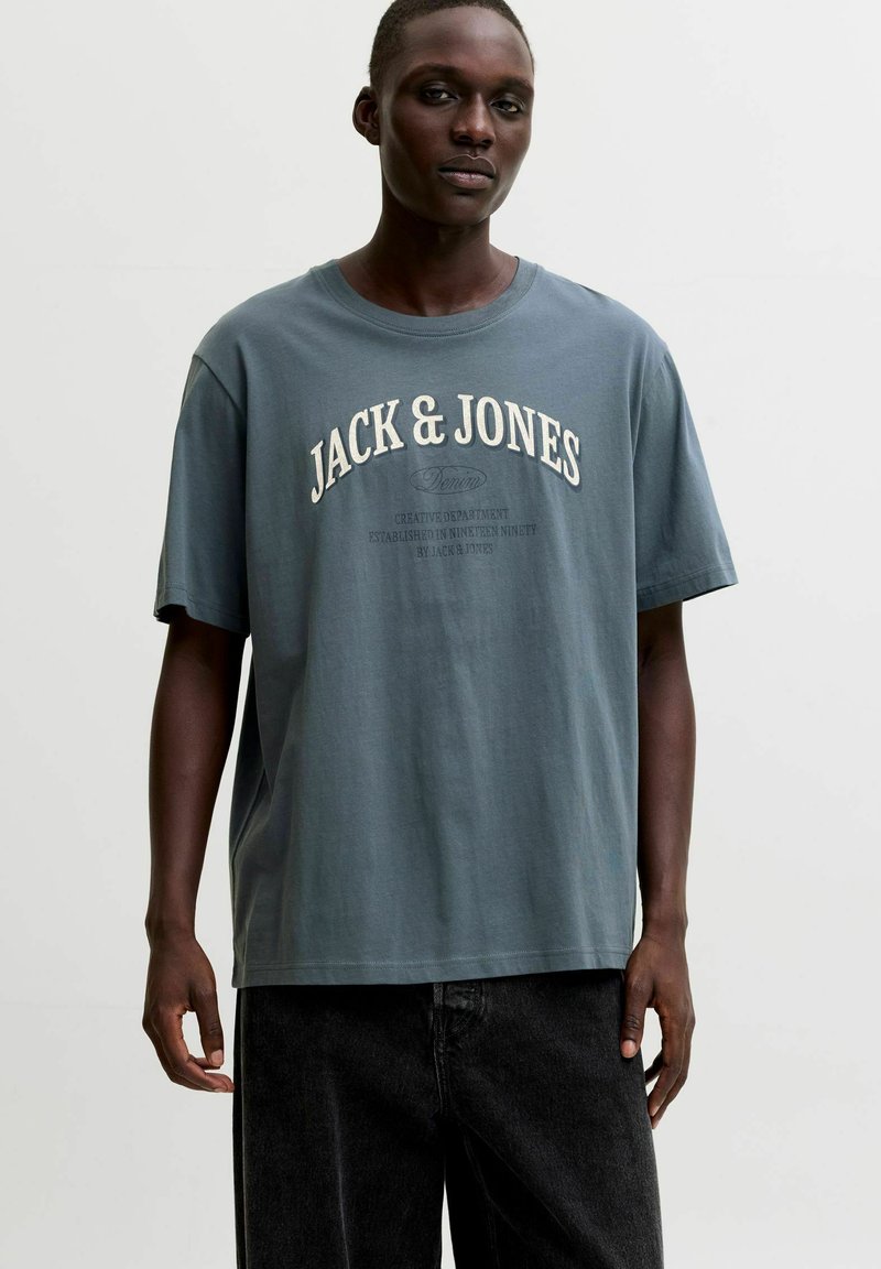 Άντρας που φοράει μπλε φαρδιά μπλουζάκι Jack & Jones και μαύρο παντελόνι, στέκεται μπροστά σε απλό ανοιχτό φόντο.
