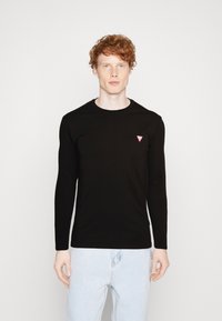 Langermet svart bomullsskjorte med rund hals og liten rød trekantlogo på venstre bryst. Brukt sammen med lyseblå denimjeans.