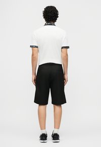 Άνδρας που στέκεται με την πλάτη γυρισμένη, φοράει λευκό πουκάμισο με κοντά μανίκια και μαύρη διακόσμηση κειμένου και μαύρα κοντά σορτς μέχρι το γόνατο με μαύρα αθλητικά παπούτσια.