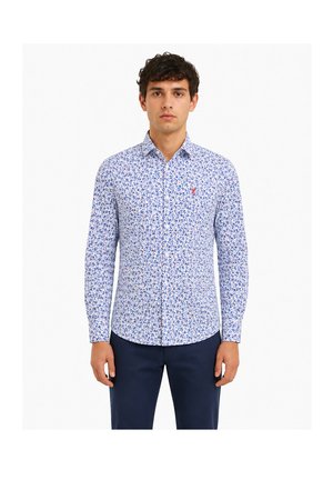 Homme portant une chemise blanche à manches longues avec de petits motifs floraux bleus et un pantalon bleu foncé, se tenant debout face à l'avant, les bras le long du corps.
