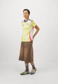 Tröja i gult och vitt med rosa detaljer, med rund halsringning, korta ärmar, laglogotyp, i kombination med bruna vida shorts och gröna sneakers.