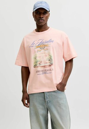 Mand iført lyserød T-shirt med fransk tekst og haveillustration, lyseblå kasket og falmede blå jeans, stående med den ene hånd i lommen.