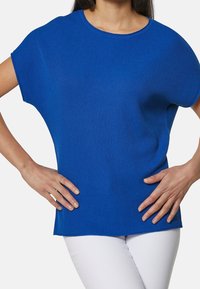 Niebieski prążkowany top z krótkim rękawem, okrągłym dekoltem oraz luźnym krojem. Materiał ma miękką fakturę; gładkie wykończenie. Noszony z białymi spodniami.