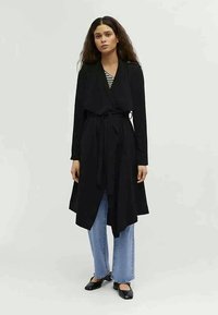 Μαύρο παλτό με μακριά μανίκια, που κλείνει σε τύπο περιτυλίγματος με ζωνάκι στη μέση, με κρεμαστή μπροστινή πλευρά και μαλακή υφή. Συνδυάζεται με μπλε φαρδιά τζιν και μαύρες πλατφόρμες. Ορατές κάθετες ρίγες.