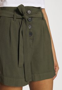 Gonna verde oliva alta in vita con bottoni neri frontali e una cintura in tessuto annodata, indossata con una maglietta bianca, mostrando una mano di lato.