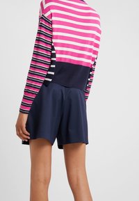 Personne portant un pull rayé rose, bleu marine et blanc, avec un short taille haute bleu marine, debout devant un fond clair uni.