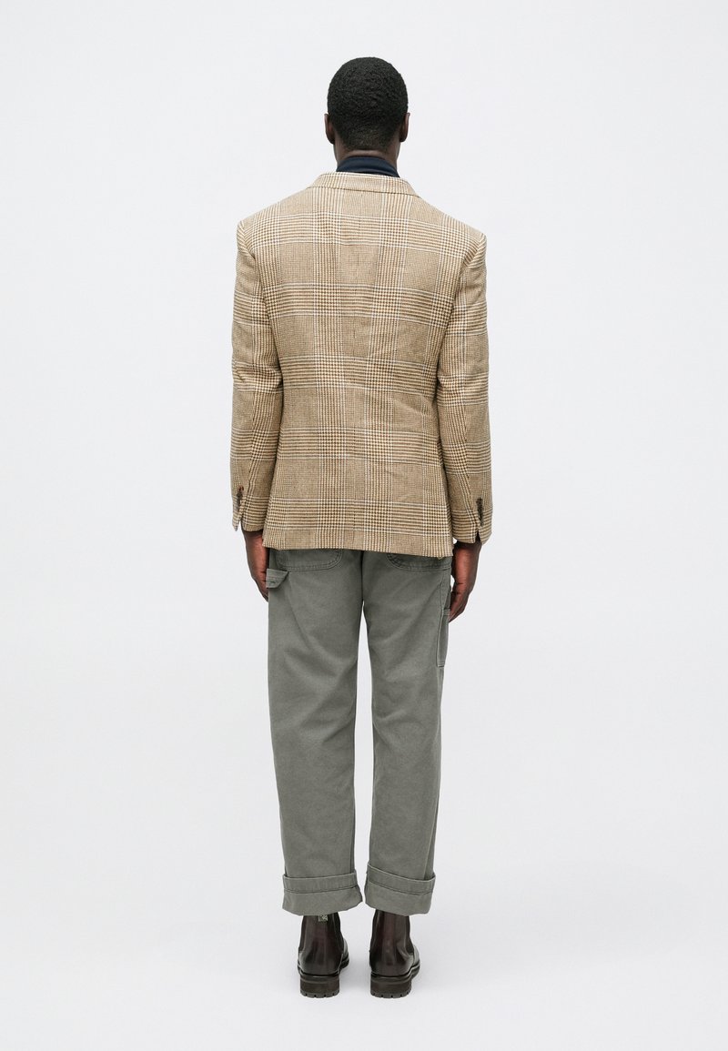 Brown checkered blazer made of textured fabric, paired with olive green cargo pants, and dark brown ankle boots, viewed from the back.