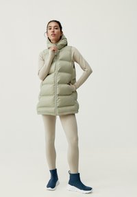 Ljusgrön pufferväst med huva, featuring en dragkedja och framträdande bafflar. Buren över en beige topp och kombinerad med marinblå ankelboots.